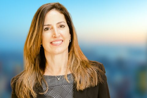 A woman with long brown hair wearing a black blazer and patterned top stands in front of a blurred outdoor background.