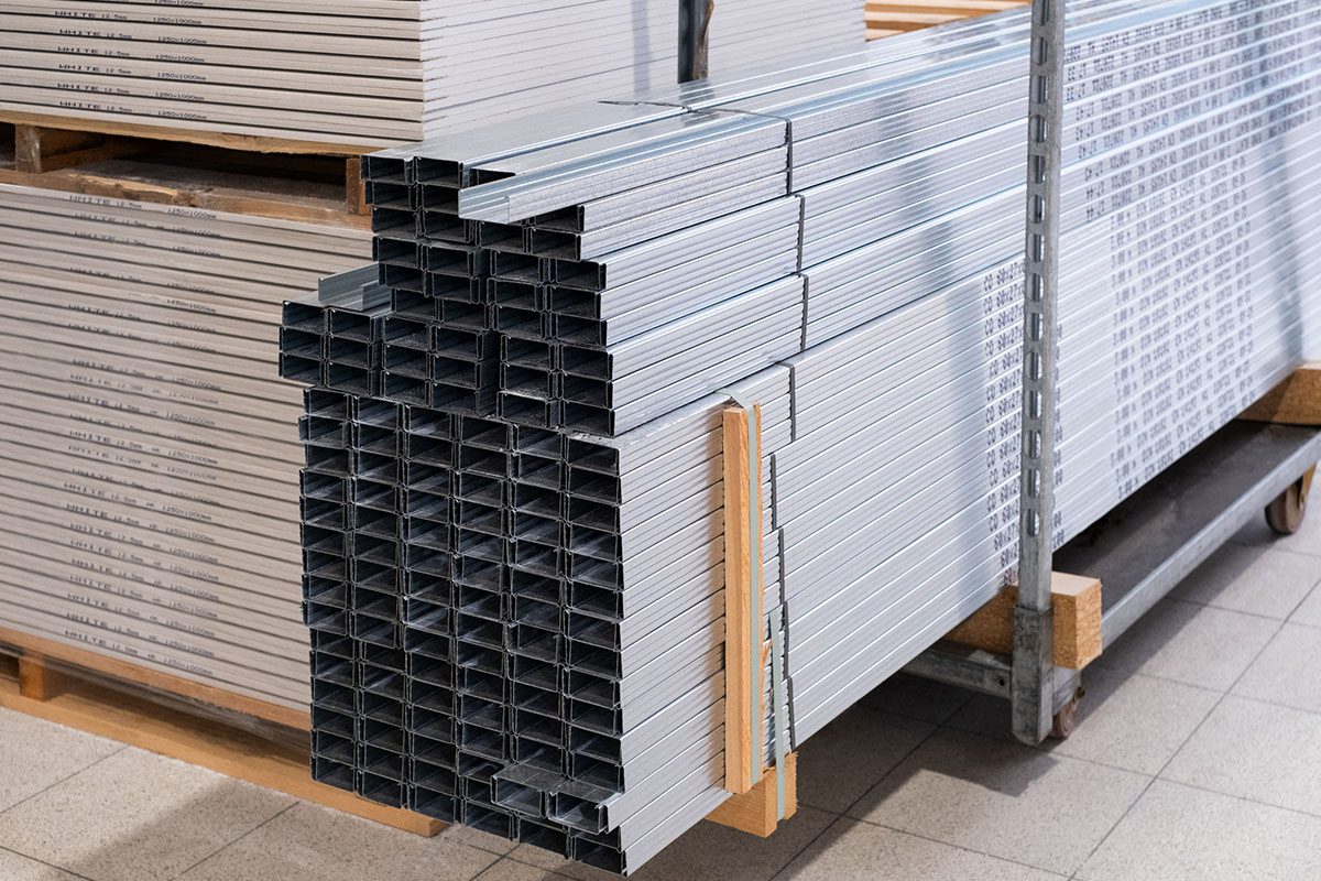 Stack of rectangular metal profiles arranged on a pallet in a warehouse, with additional metal sheets stored on shelves in the background.