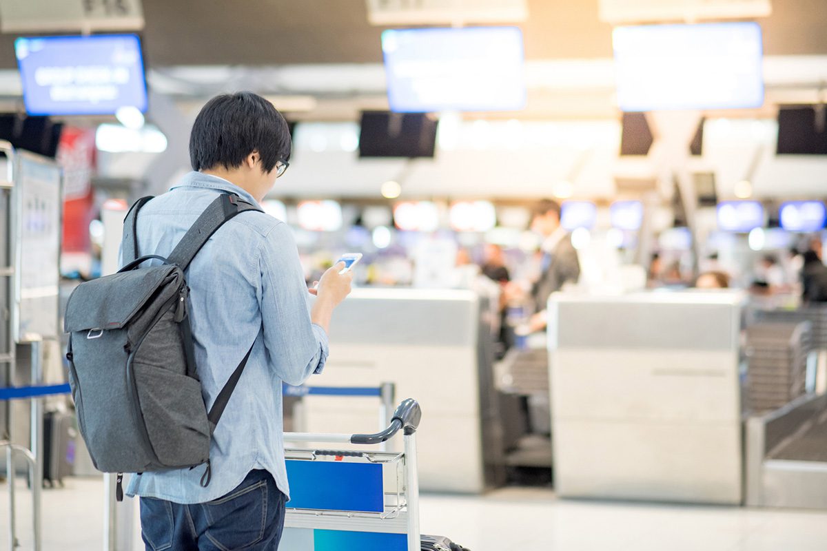 A person with a backpack stands at an airport check-in counter, looking at their phone while pushing a luggage cart.