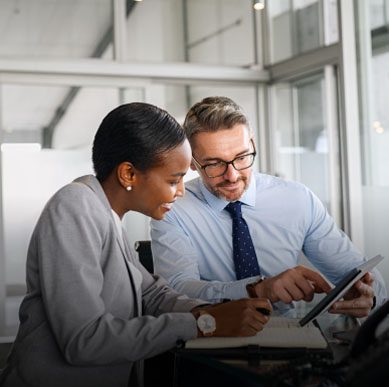 Two professionals in business attire sit at a desk, looking at a tablet and discussing information in a modern office setting.
