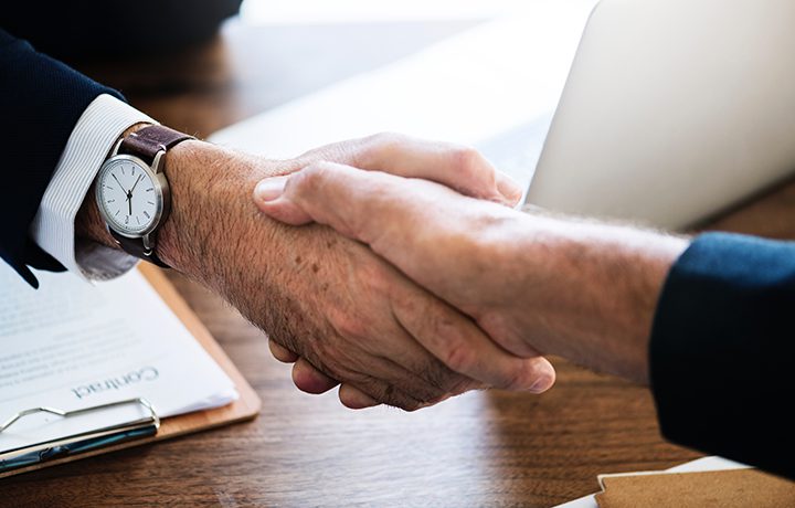 Two people in business attire shaking hands over a desk with documents and a laptop.