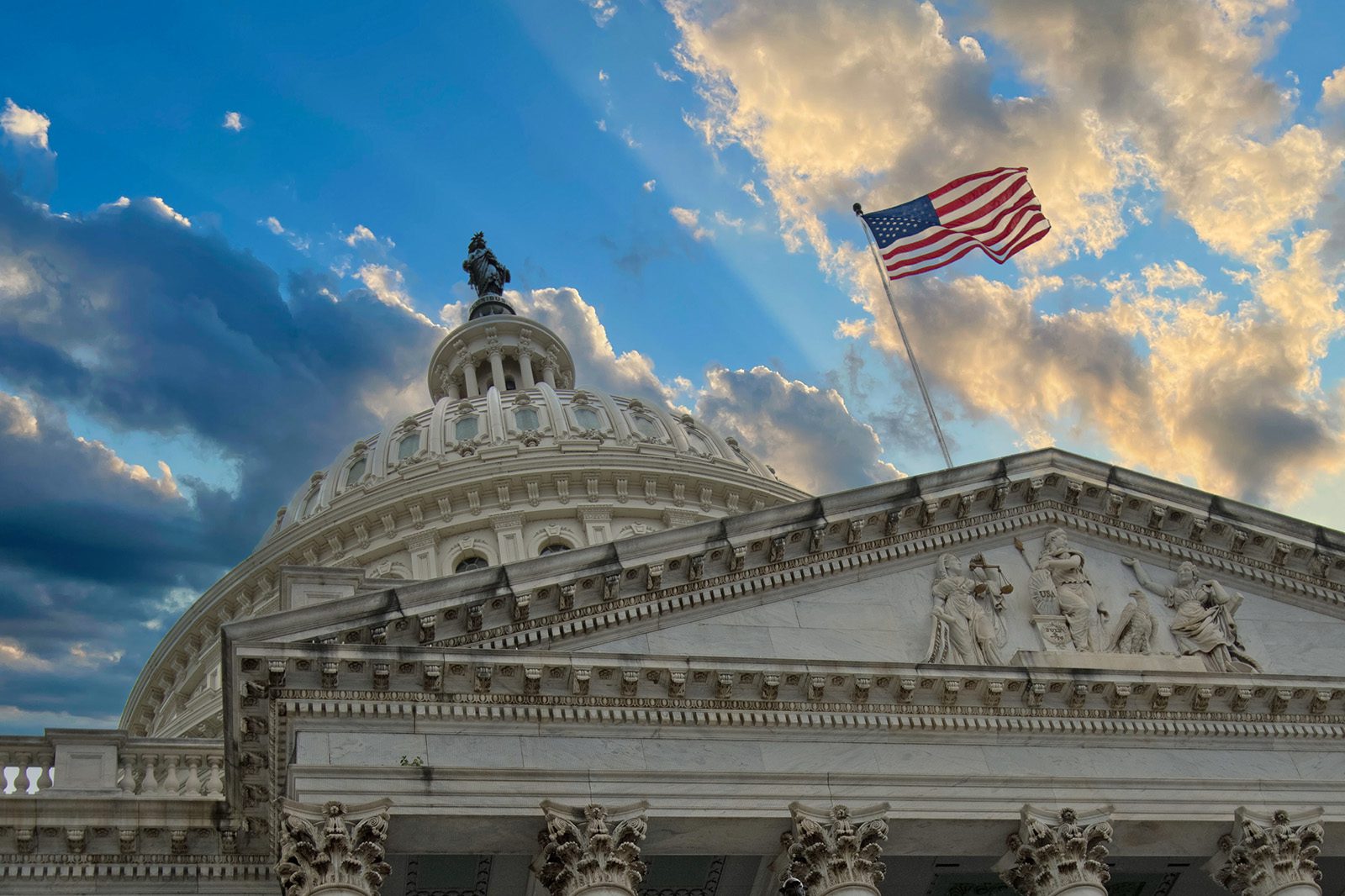 The U.S. Capitol building dome with a waving American flag in front and a partly cloudy sky at sunset in the background.