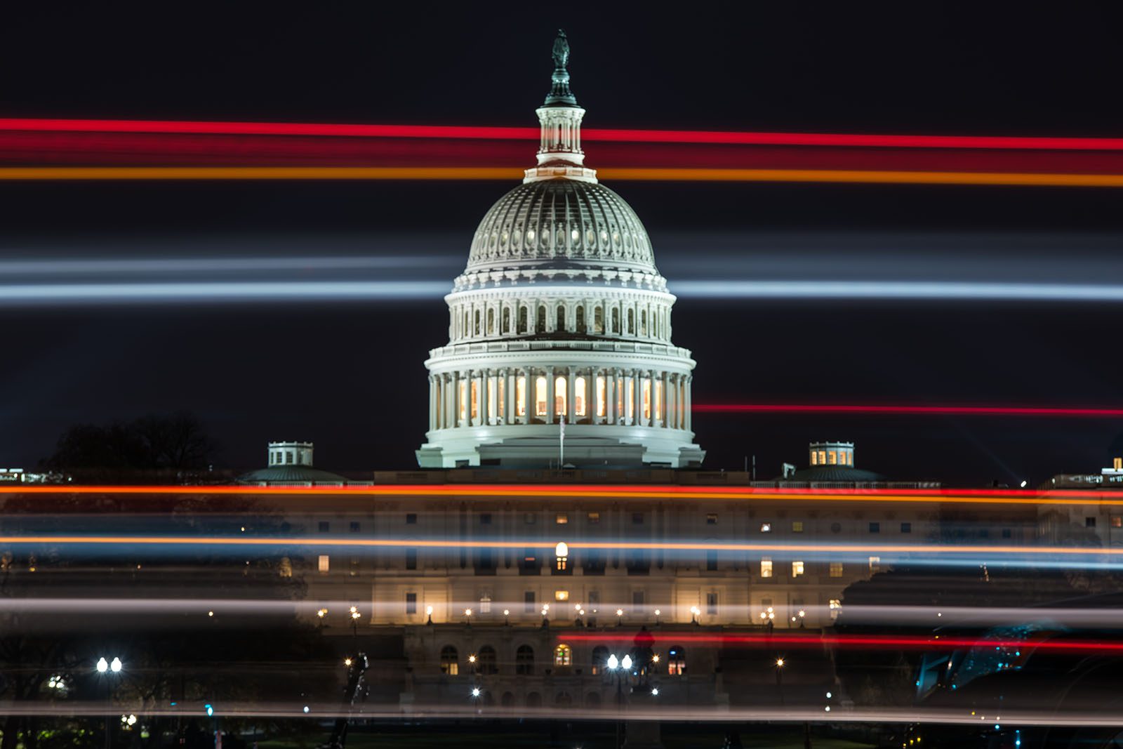 The U.S. Capitol building at night, illuminated, with blurred horizontal light trails from passing vehicles in the foreground.