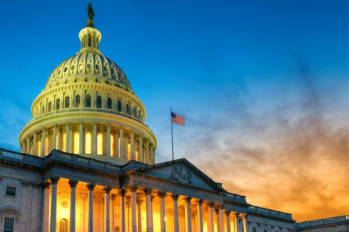 The U.S. Capitol building illuminated at dusk, with the American flag flying and a colorful sunset sky in the background.
