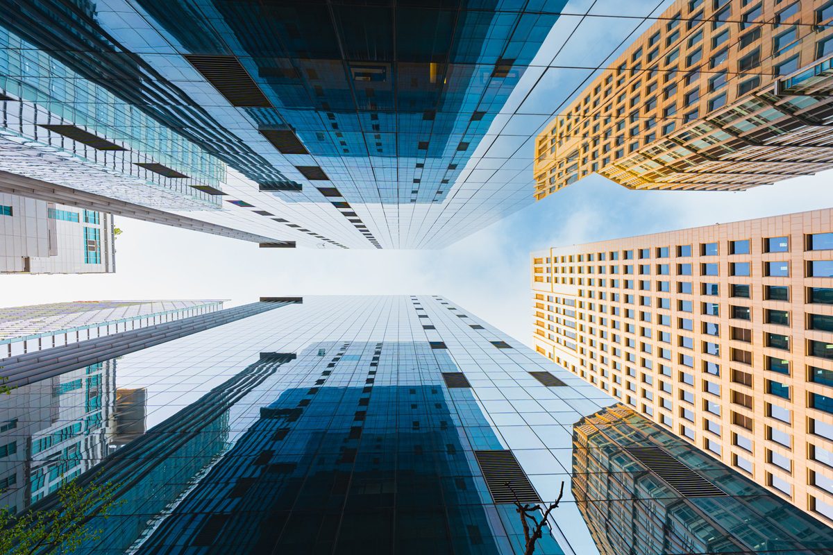 View looking up at tall modern skyscrapers with glass and steel facades against a blue sky, creating a symmetrical, geometric effect.