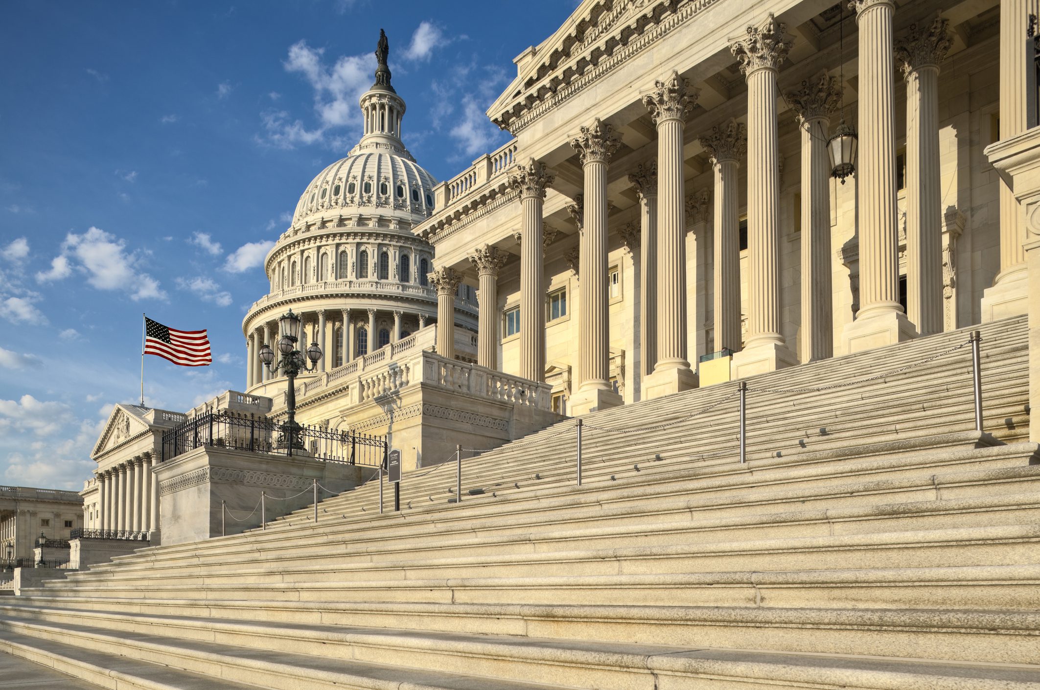 The U.S. Capitol building with the American flag flying, seen from the steps under a partly cloudy sky.
