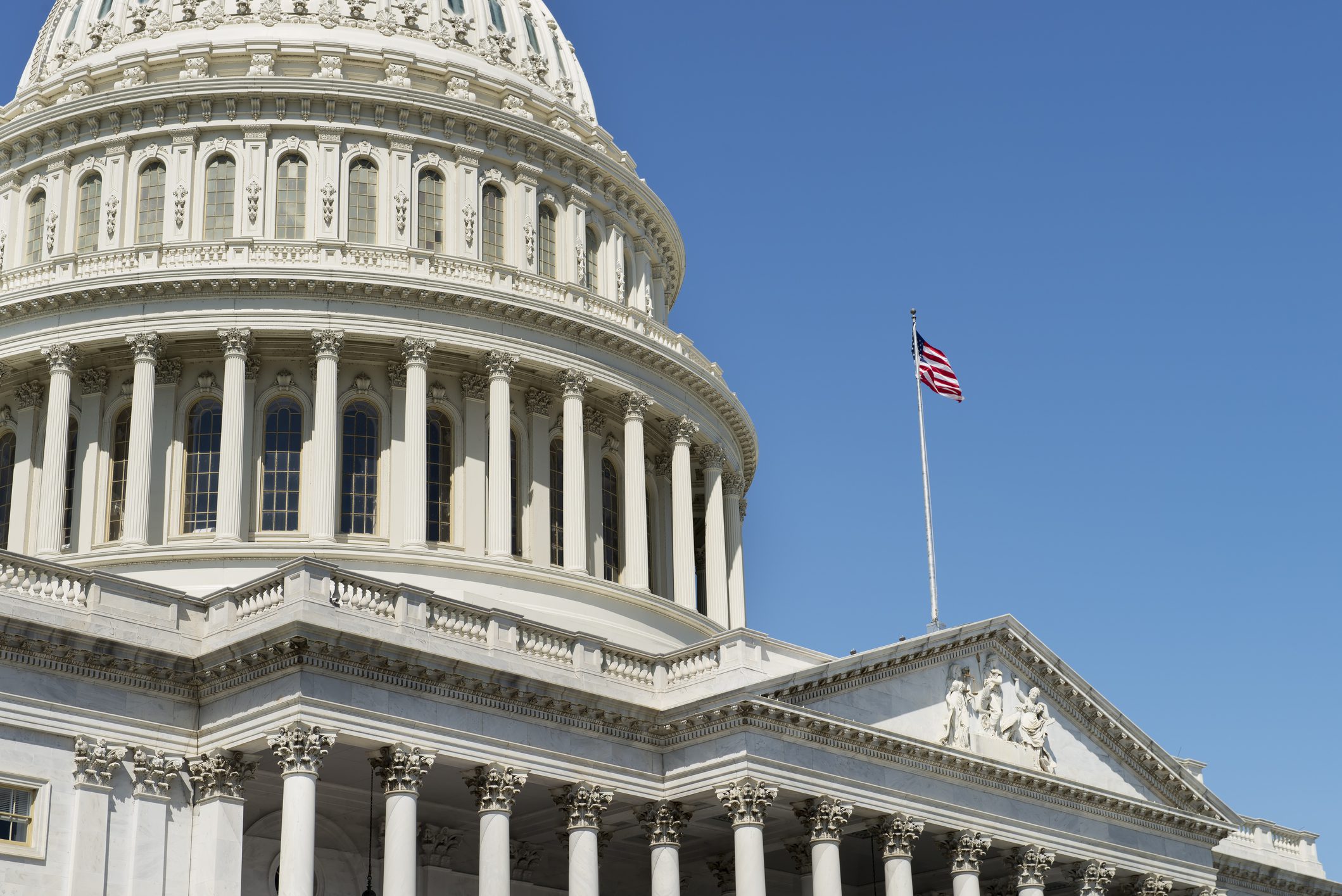 A close-up of the United States Capitol building dome with an American flag on a flagpole against a clear blue sky.