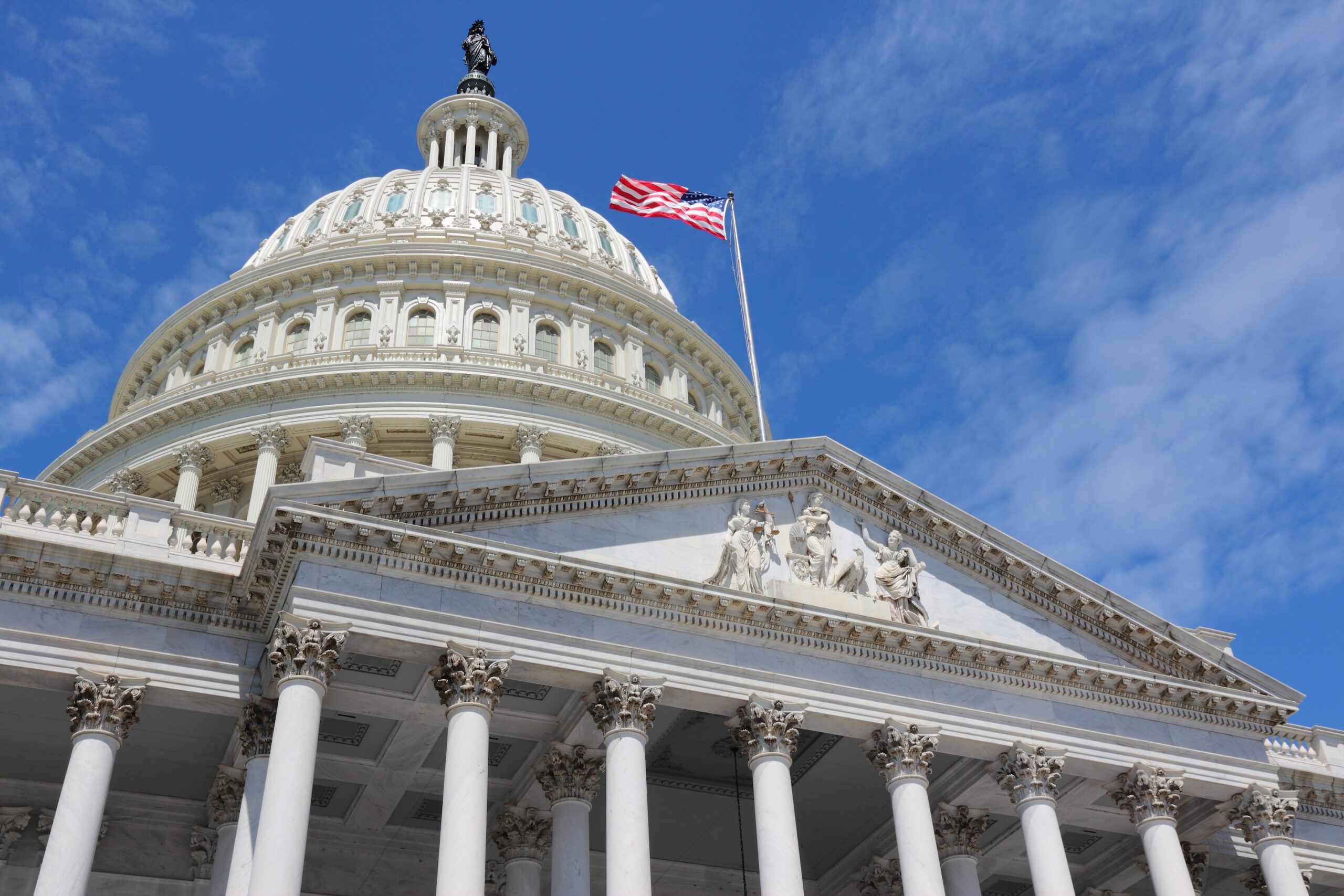 The United States Capitol building with a U.S. flag flying, viewed from below against a blue sky with scattered clouds.