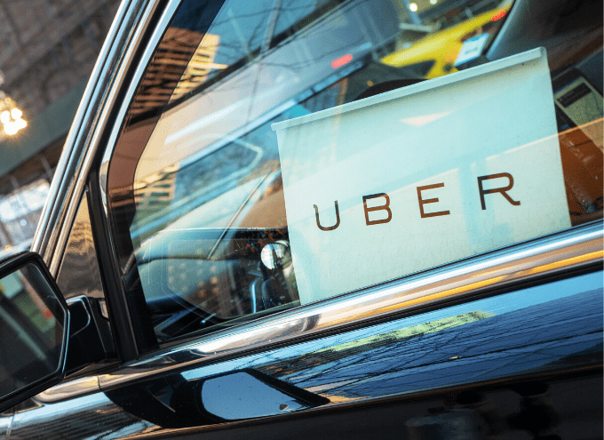 A car window with an Uber sign displayed on the dashboard, reflecting city buildings and street activity outside.