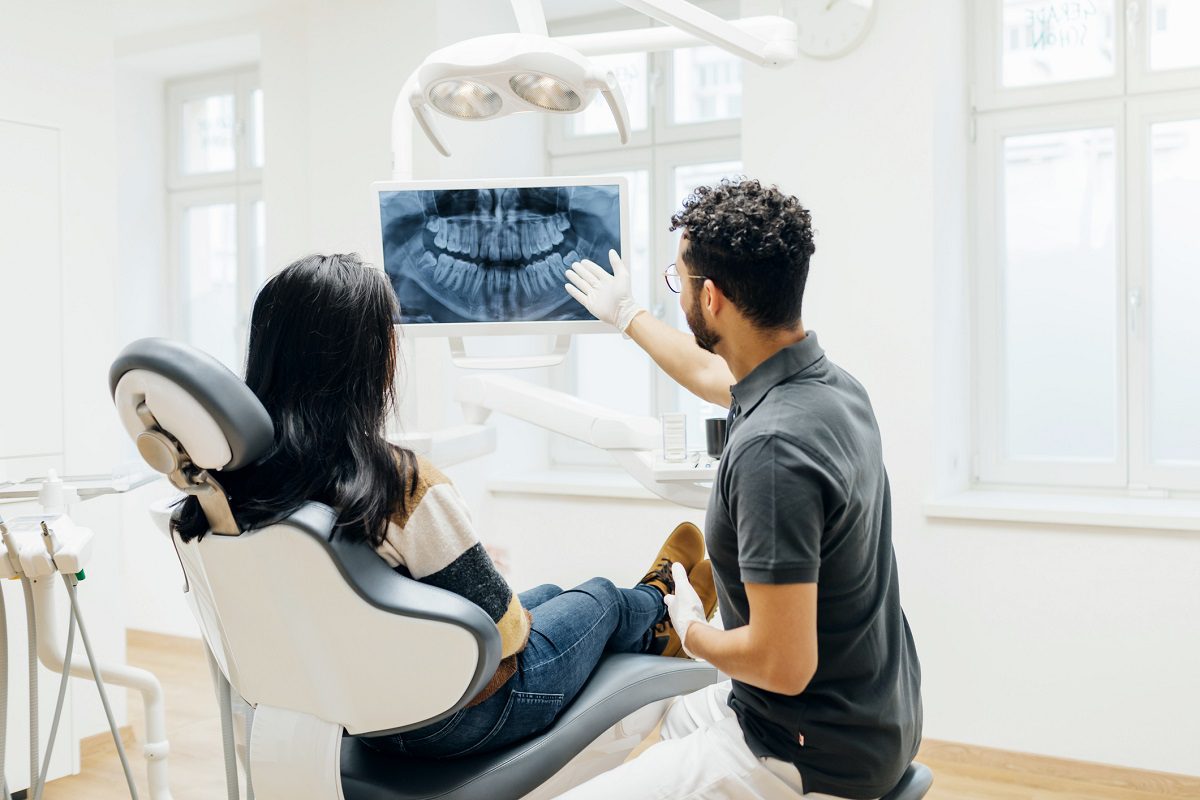 A dentist shows a dental X-ray on a screen to a patient sitting in a dental chair in a bright, modern clinic.