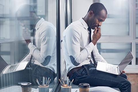Man in business attire sits on a desk with a laptop, appearing thoughtful, with his reflection visible in a glass wall beside him.