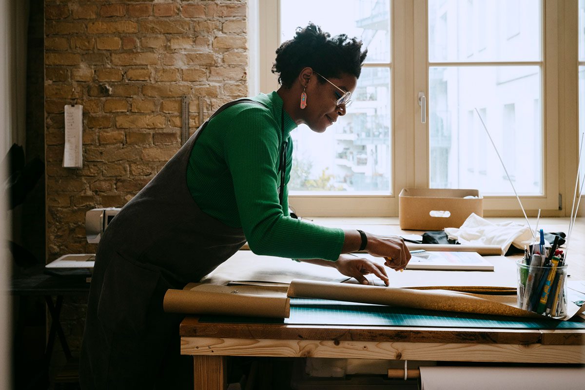 A person wearing glasses and an apron works with paper and tools on a table in a well-lit room with brick walls and large windows.