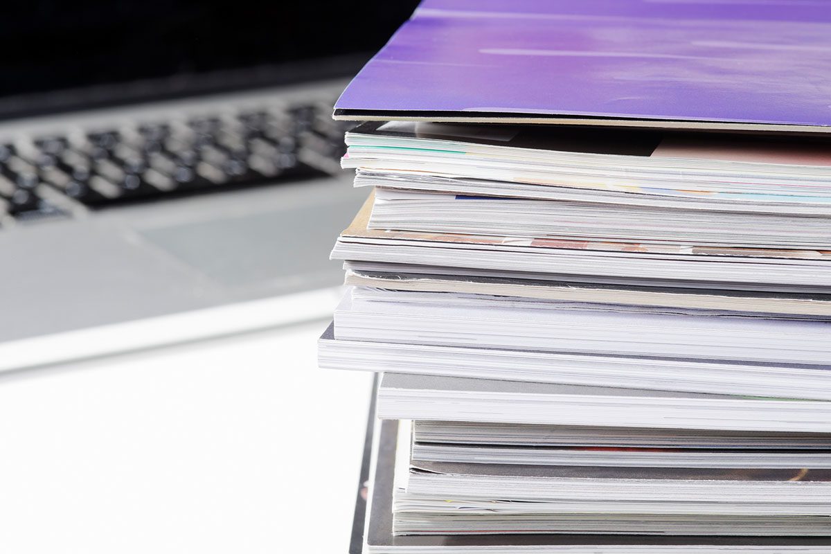 A stack of magazines and documents sits on a desk next to an open laptop.
