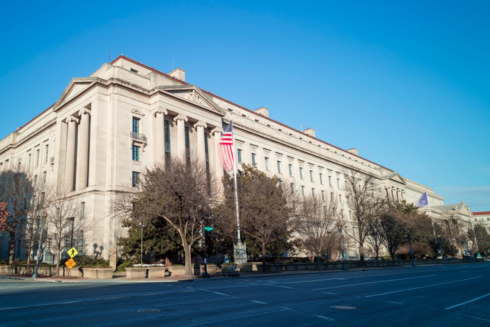 A large government building with columns and a US flag in front, set on a nearly empty street with leafless trees under a clear blue sky.