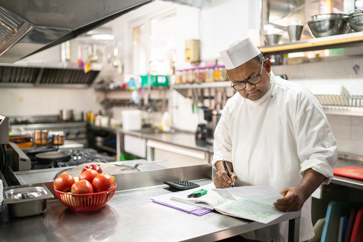 A chef in a white uniform and hat writes in a notebook in a commercial kitchen, with tomatoes in a bowl and kitchen equipment in the background.