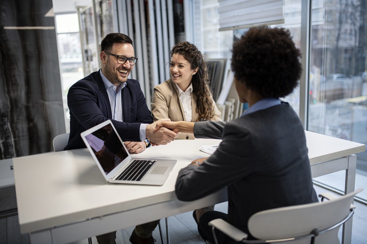 Three people in business attire sit at a table in an office; two are shaking hands while the third person observes. A laptop is open on the table.