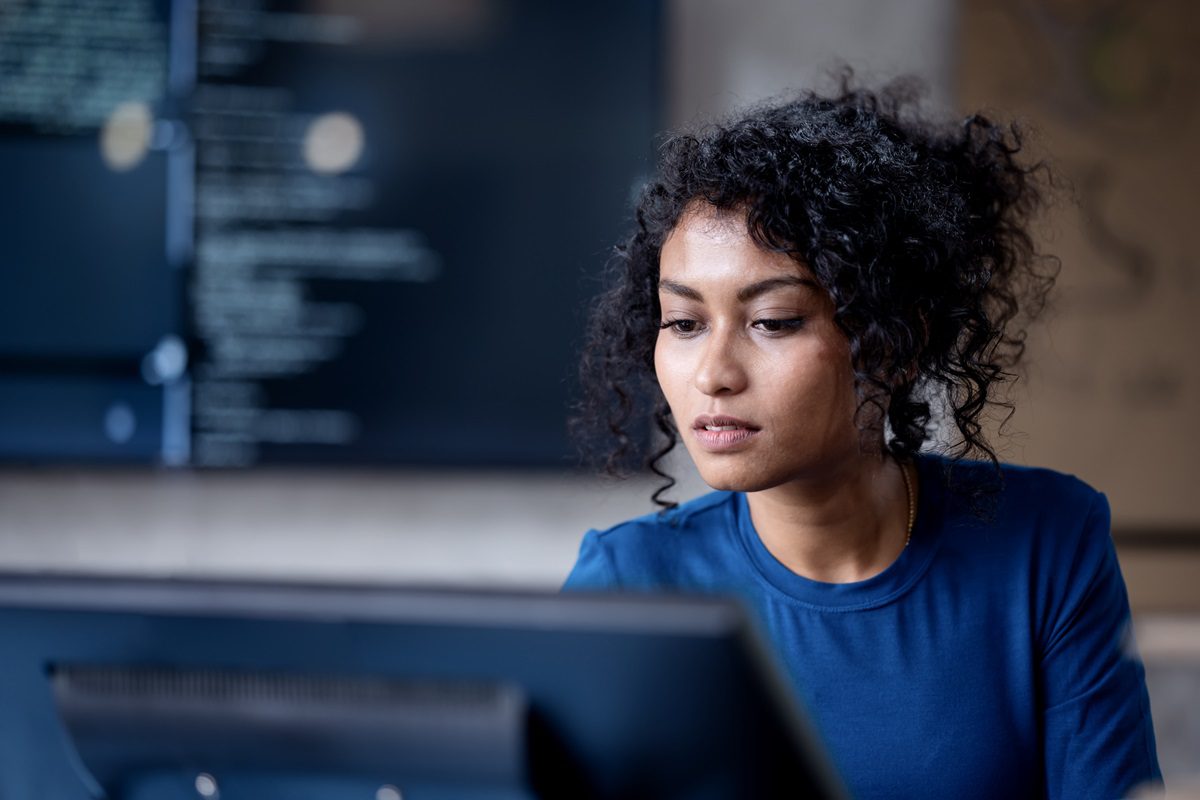 A woman with curly hair wearing a blue shirt looks at a computer monitor, with code visible on a screen in the background.