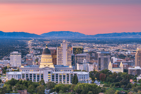 Downtown cityscape at sunset featuring a domed capitol building in the foreground, high-rise buildings, and mountains in the background.