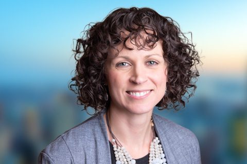 Woman with dark curly hair and a silver blazer smiles in front of a bright window with a blurred city view