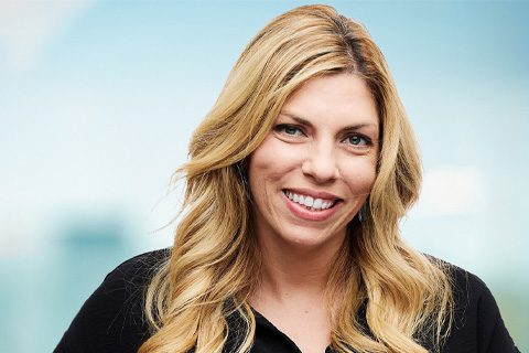 A woman with long, wavy blonde hair and a black top smiles at the camera against a blurred blue background.