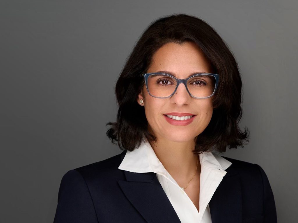 A woman with dark hair and glasses, wearing a white collared shirt and dark blazer, poses for a professional headshot against a plain gray background.