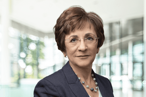 A woman with short brown hair, glasses, and a blue suit jacket with a beaded necklace stands in a bright, modern office setting.