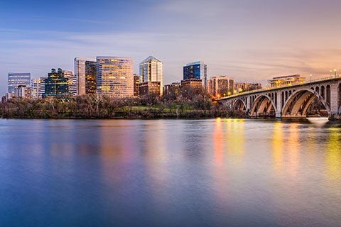 A cityscape with modern buildings and a bridge over a calm river at sunset, with reflections visible on the water.