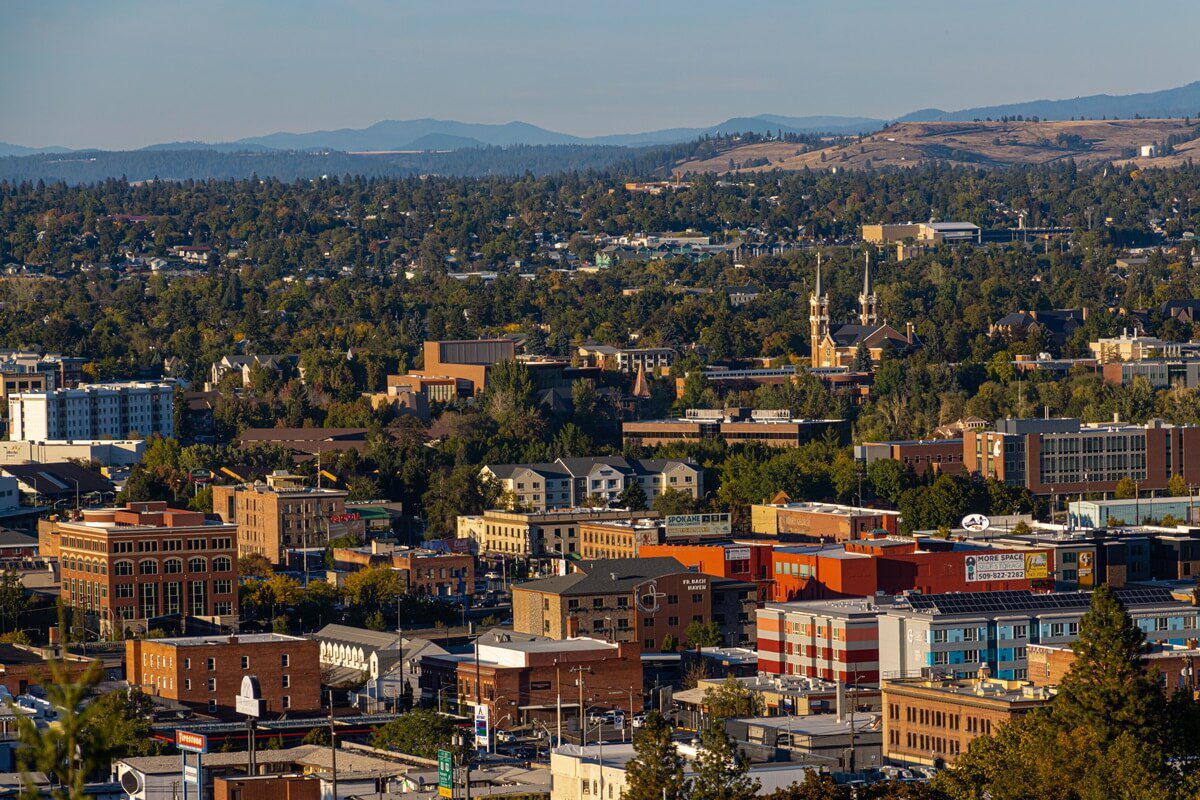 Aerial view of a city with commercial and residential buildings, tree-lined streets, and hills in the background under a clear sky.