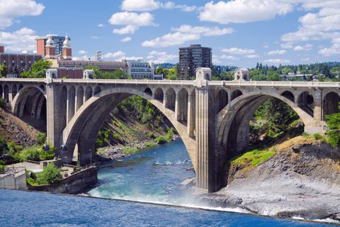A large stone arch bridge spans a river with buildings and trees in the background under a partly cloudy sky.