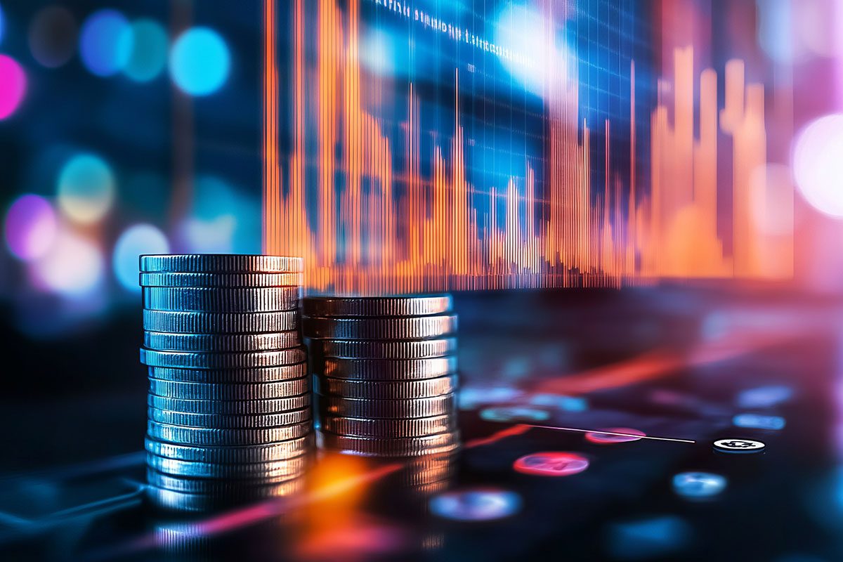 Stacks of coins in the foreground with colorful digital financial graphs and charts displayed in the blurred background.