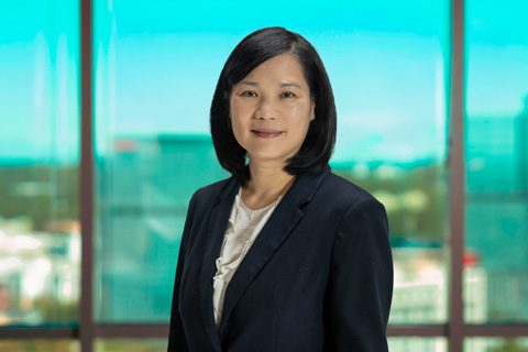 A woman with shoulder-length black hair wearing a dark blazer and white blouse stands indoors in front of large windows with a cityscape in the background.