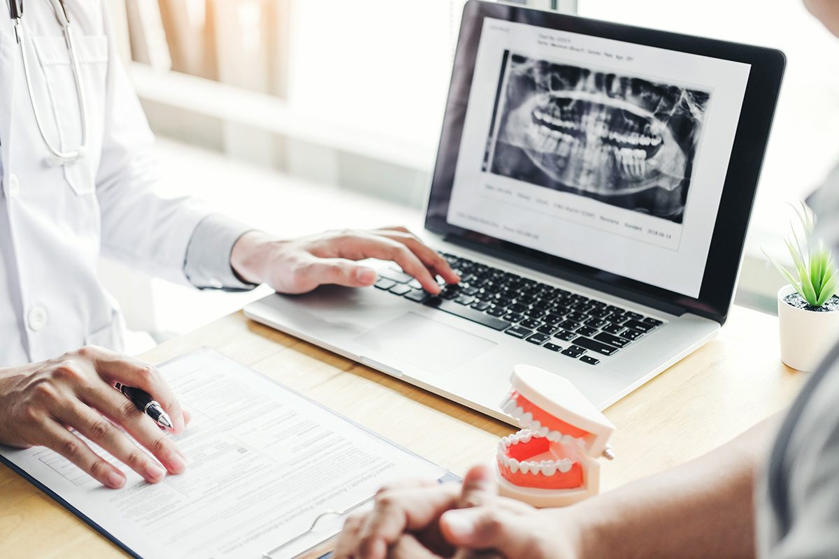 A dentist reviews a dental x-ray on a laptop with a patient, while pointing at a form on a clipboard; a dental model sits on the desk.