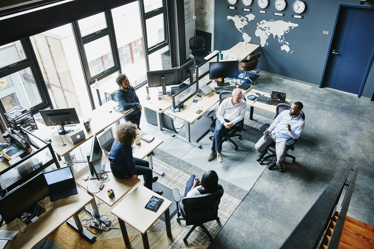 Five people sit in a modern office with multiple desks, computers, and wall clocks showing different time zones, engaged in a group discussion.