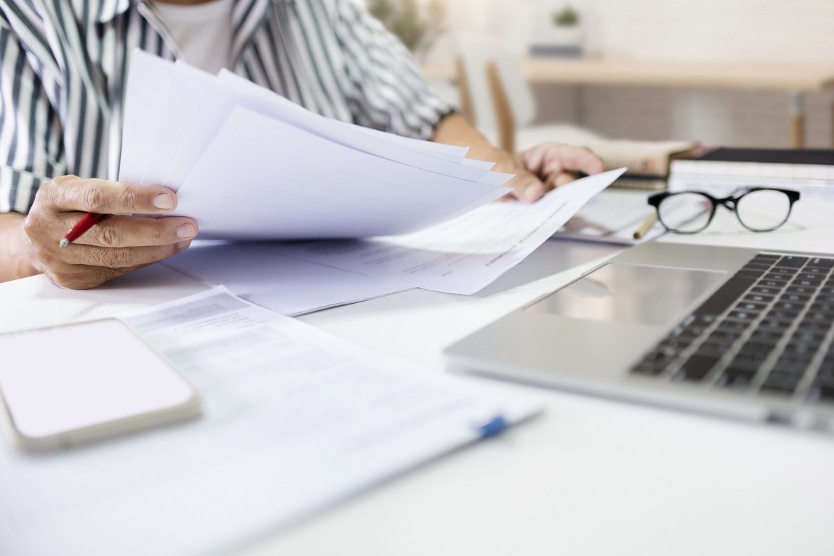 Person holding documents at a desk with a laptop, smartphone, glasses, and paperwork, suggesting an office or work-from-home environment.