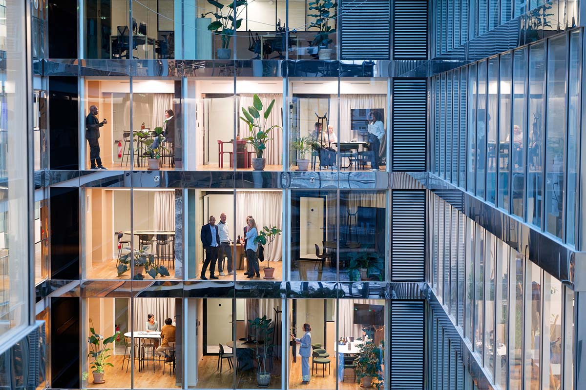 People are seen working, conversing, and walking in a modern multi-story office building with glass walls and indoor plants.
