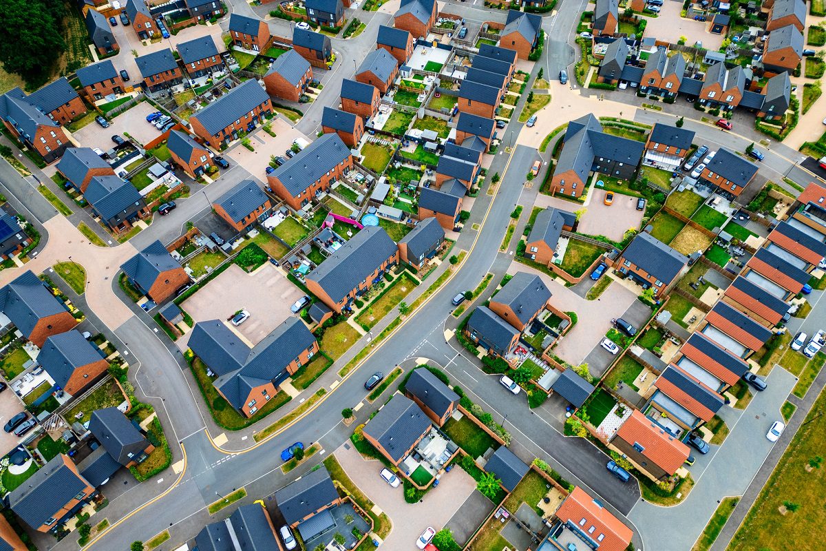 Aerial view of a suburban neighborhood with rows of houses, driveways, parked cars, and neatly arranged lawns separated by winding streets.