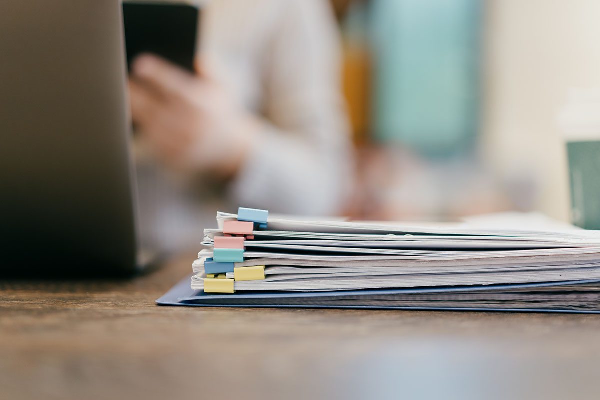 A stack of documents with colorful paper clips sits on a wooden desk, with a laptop and a takeaway coffee cup visible in the blurred background.
