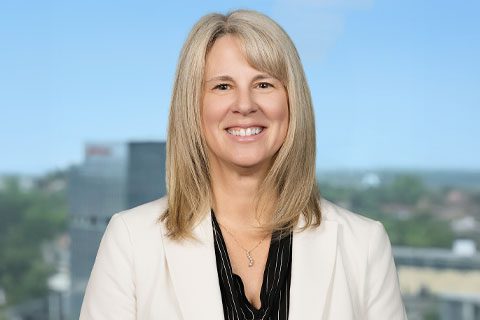 Blonde woman in a white blazer smiles in front of a bright window with a blurred city view.