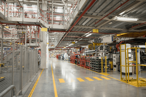 Interior of a modern industrial factory showing machinery, metal structures, storage racks, and safety markings on the polished floor.