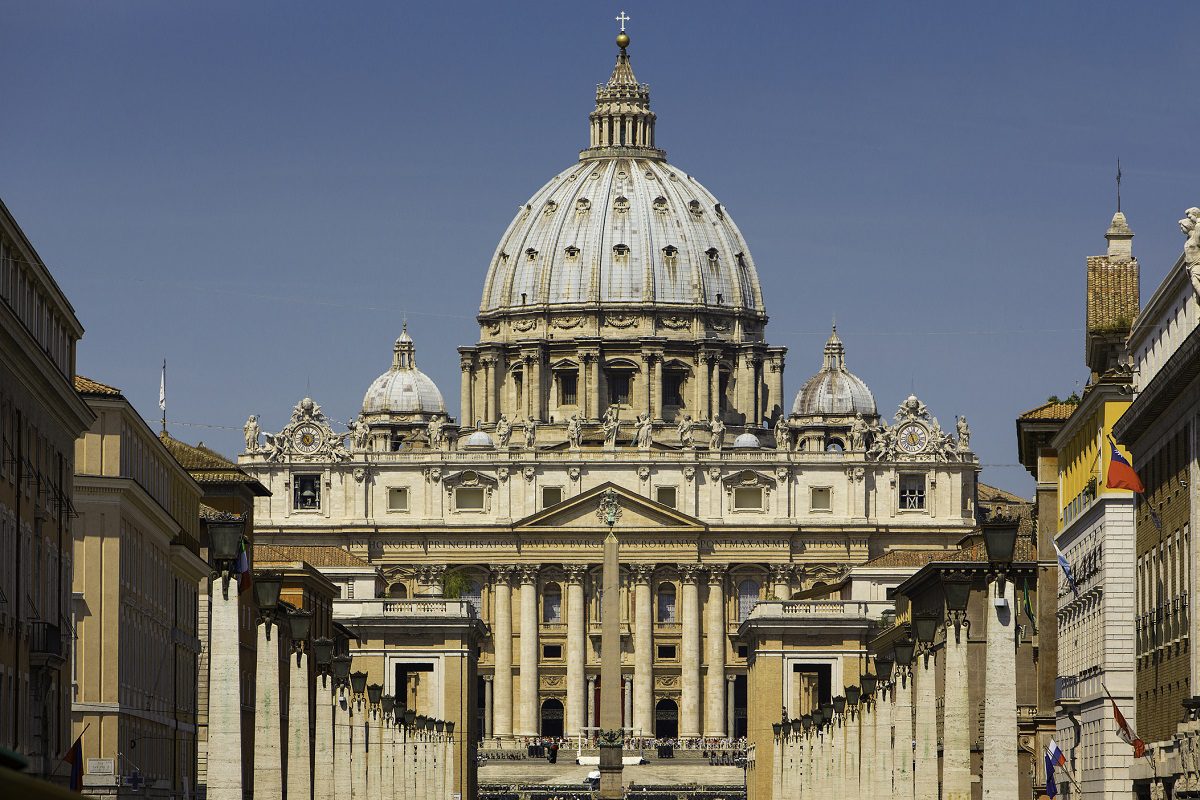 St. Peters Basilica in Vatican City, featuring its large central dome and symmetrical façade, viewed from the street with columns lining the approach.