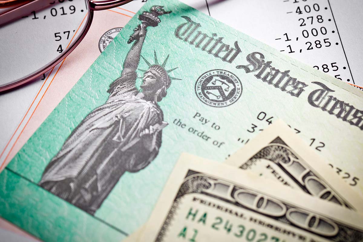 Close-up of a U.S. Treasury check and dollar bills on top of financial documents, with eyeglasses partially visible in the background.