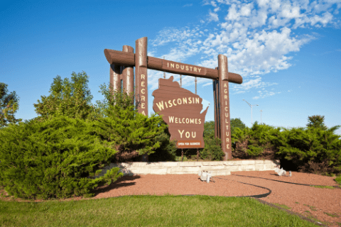 Large wooden sign reading Wisconsin Welcomes You surrounded by greenery, with Industry, Recreation, and Agriculture written on overhead beams.