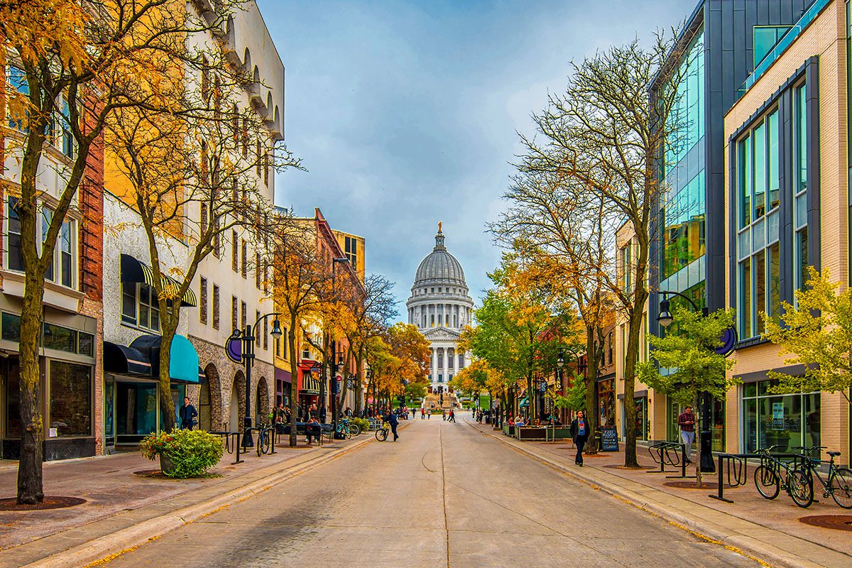 A wide street lined with buildings and trees leads towards a large domed capitol building under a cloudy sky. People walk and ride bikes along the sidewalks.