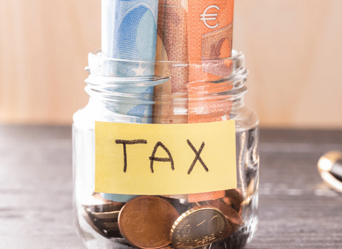 A glass jar labeled TAX contains euro banknotes and coins, placed on a wooden surface.
