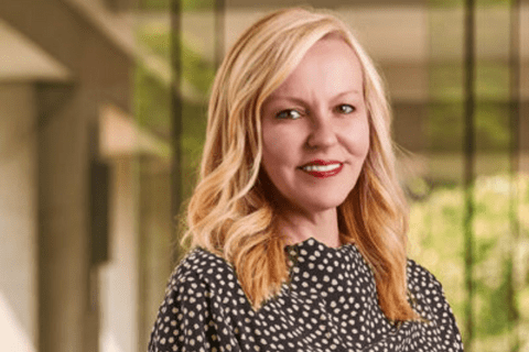 A woman with shoulder-length blonde hair, wearing a black and white polka dot blouse, stands indoors with blurred green scenery visible through large windows in the background.