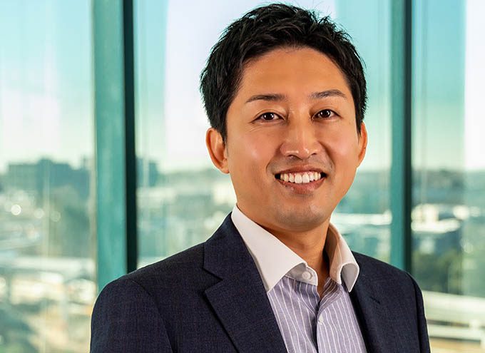 A man in a suit and collared shirt stands indoors in front of large windows with a cityscape in the background, smiling at the camera.