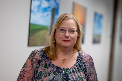Blonde woman in glasses and a floral top smiles in a blurred hallway