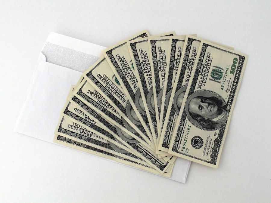 A white envelope with several one hundred dollar bills fanned out on top of it, placed on a plain white background.