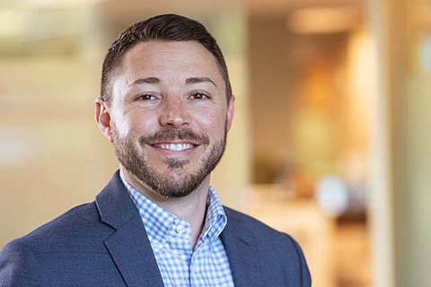 A man with short dark hair and a beard, wearing a blue suit jacket and checked shirt, smiles at the camera in an office setting.