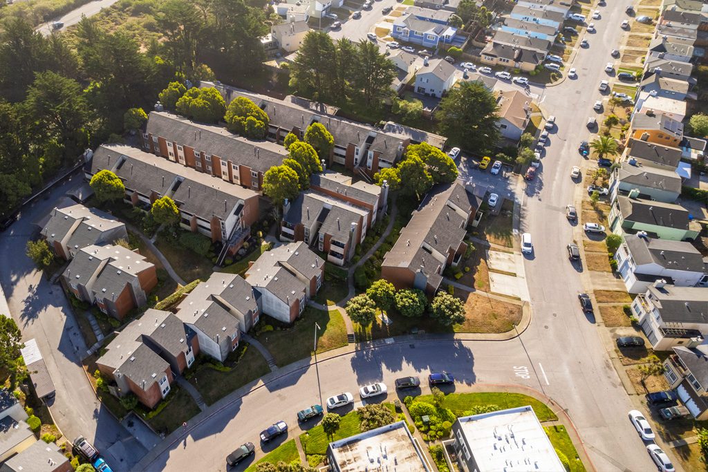 Aerial view of a residential neighborhood with apartment buildings, houses, trees, streets, and parked cars on a sunny day.